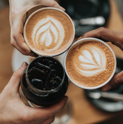 Top-down view of three hands clinking two latte cups and one iced coffee over a wooden table